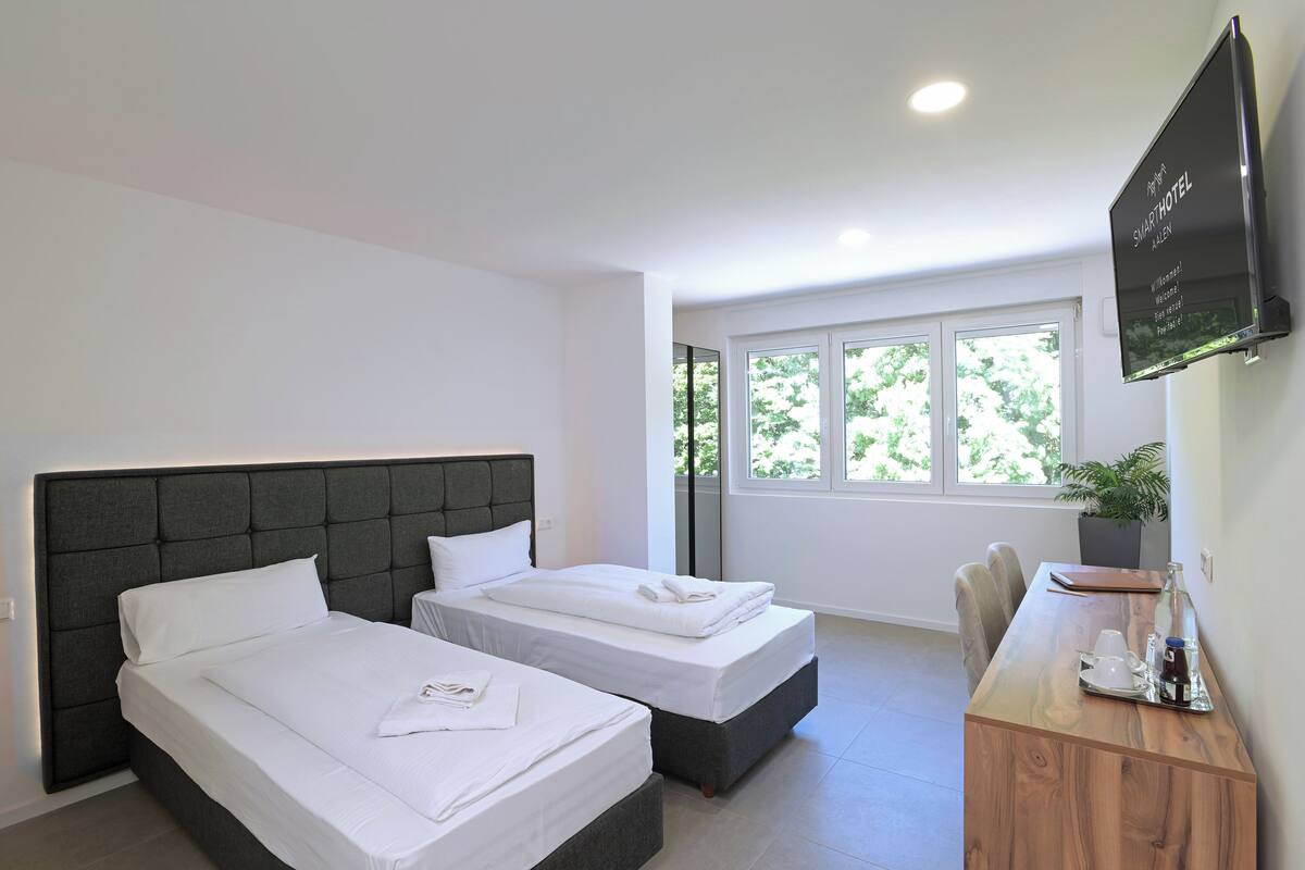 A hotel room with two beds, a desk with a chair, a TV, and a window with a view of trees.