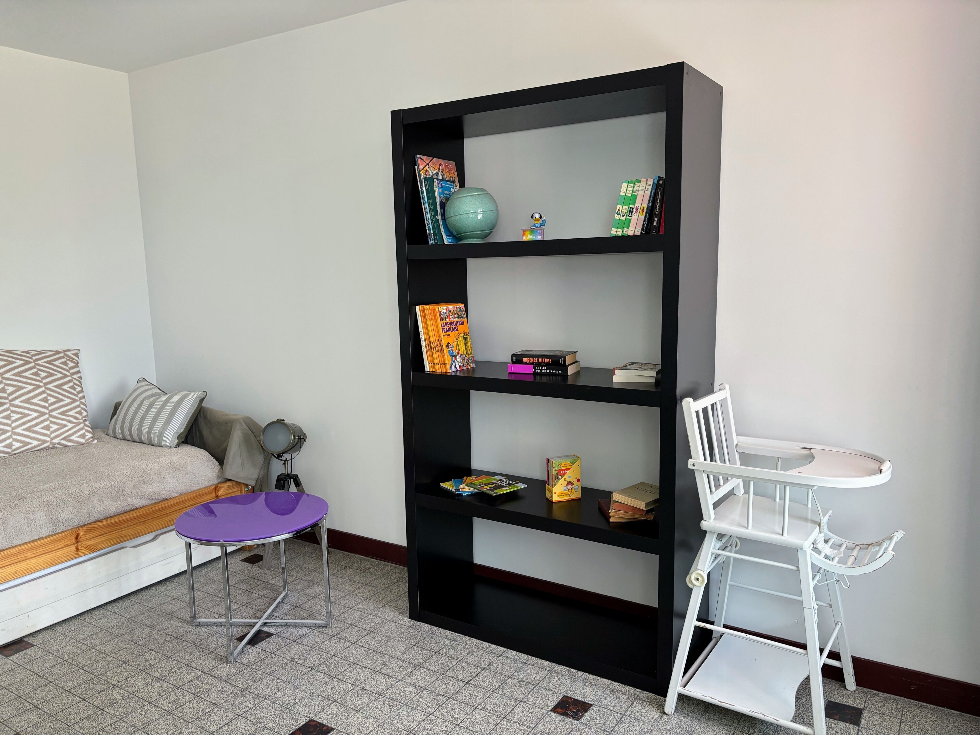 Ein Zimmer mit einem Bett, einem kleinen violetten Sessel, einem weißen Stuhl, einem schwarzen Bücherregal und einem kleinen Tisch.