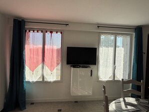 A room with a TV on a stand, a window with curtains, a chair, and a table. - City center apartment with garden, near the sea (Quimperlé)