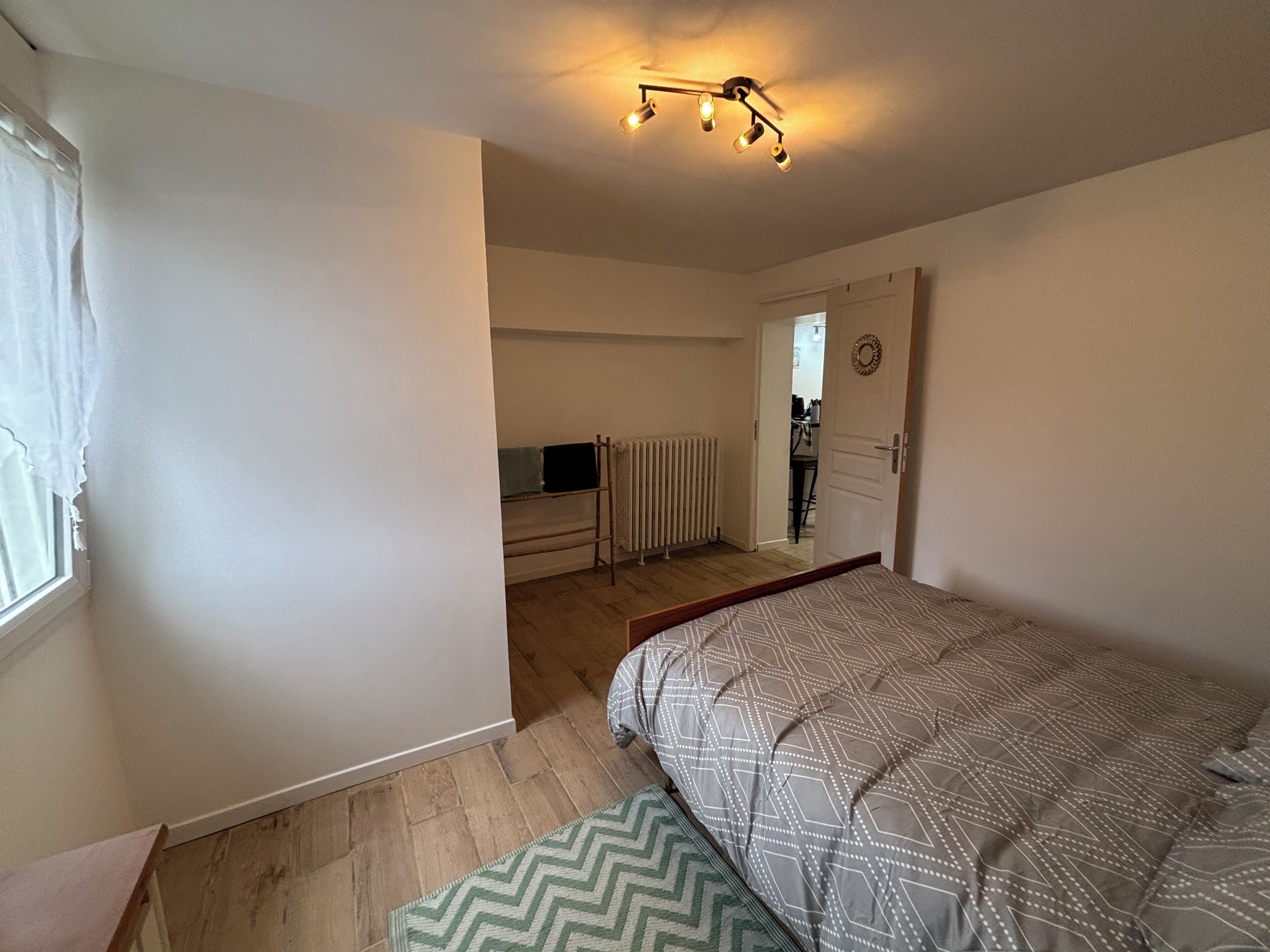 Een slaapkamer met een bed, een radiator, een televisie op een standaard en een deur naar een andere kamer.