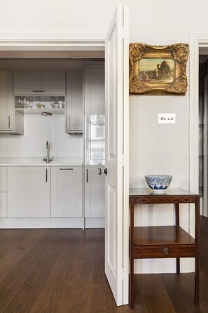 A kitchen with white cabinets, a wooden table with a bowl, and a gold-framed picture on the wall. - Chic 1-bed in Notting Hill for 3 (London)