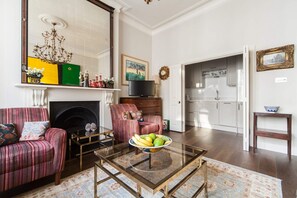 A living room with a fireplace, striped sofas, a glass coffee table, and a kitchen in the background. - Chic 1-bed in Notting Hill for 3 (London)