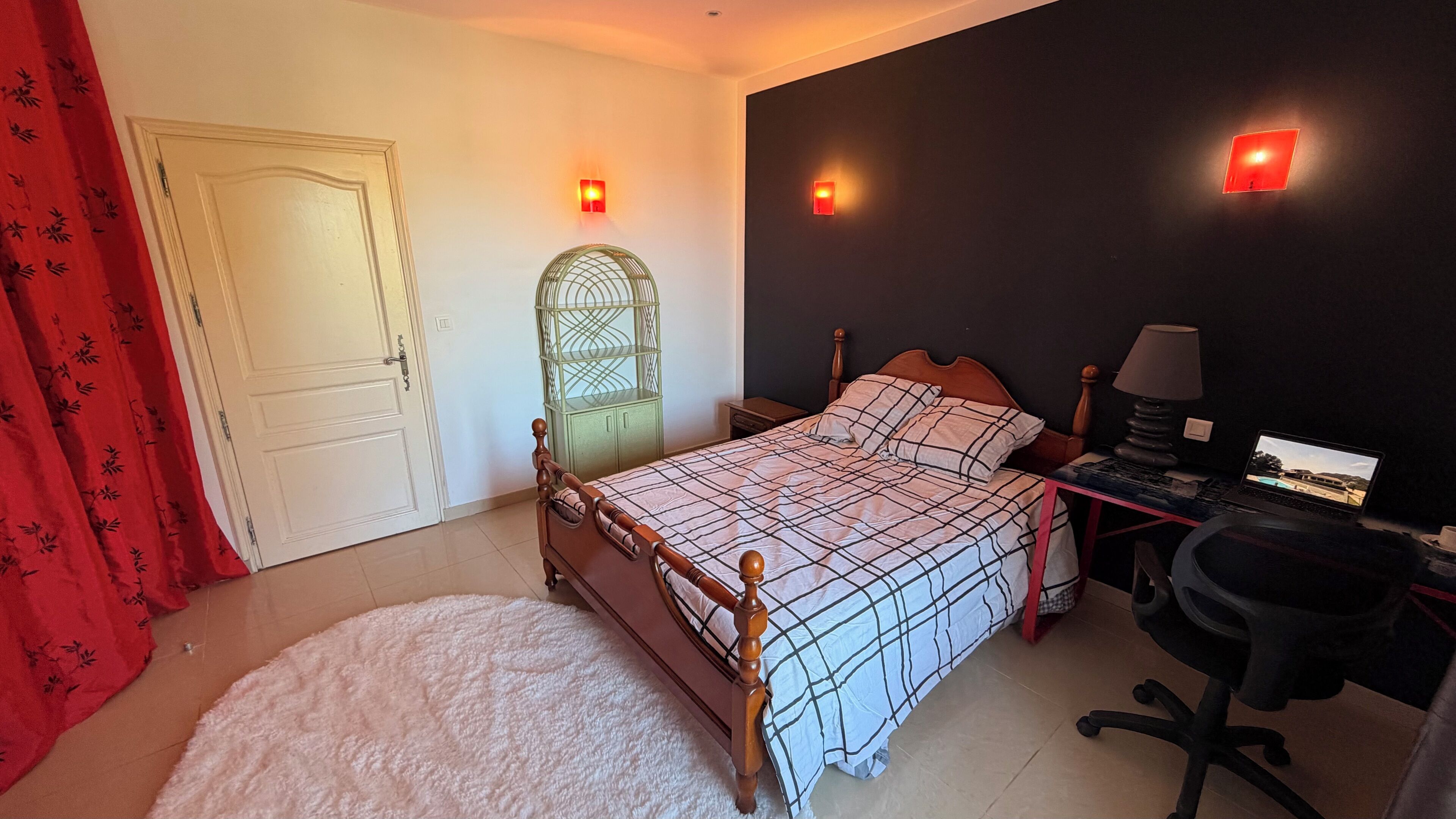 Quarto com cama, escrivaninha com laptop, cadeira, guarda-roupa e cortina vermelha.