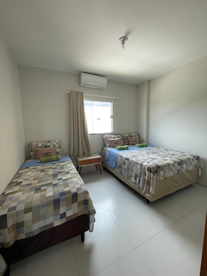 A room with two beds, a small table, a window with curtains, and an air conditioning unit.
