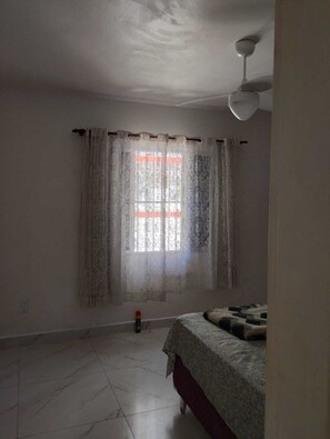 Ein Zimmer mit Bett, einem fensterbepflanzten Fenster, einem Deckenventilator und einem Fliesenboden.