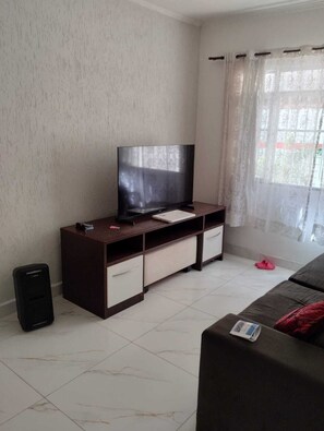 A living room with a flat-screen TV on a dark wooden TV stand, a white tiled floor, a black speaker, a red slipper, and a sofa with a magazine.