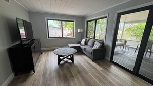 Una habitación con una televisión de pantalla plana, un sofá gris, una mesa redonda y un amplio ventanal corredero que da a un patio exterior.