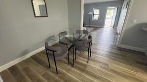 Un comedor con una mesa de vidrio y cuatro sillas, un espejo en la pared y un pasillo que conduce a otra habitación.