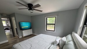 Un dormitorio con cama, ventilador de techo, televisor sobre un mueble bajo, una ventana y una puerta que da a otra habitación.