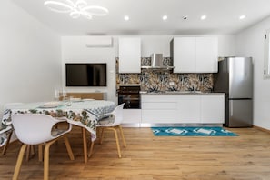 A compact kitchen with white cabinets, a stainless steel refrigerator, a flat-screen TV, and a dining area with a table and chairs.