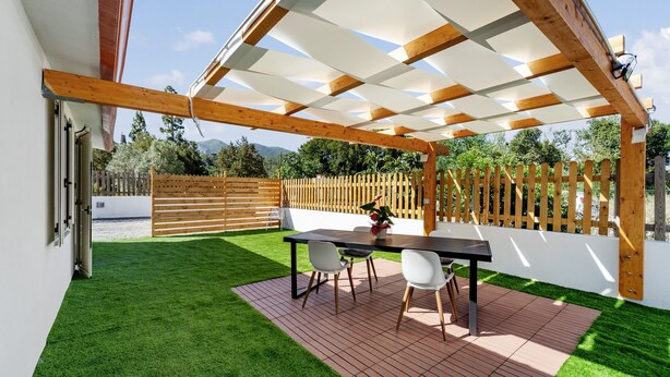 Une terrasse couverte avec une table et des chaises, entourée de gazon synthétique et d’une plateforme en bois.