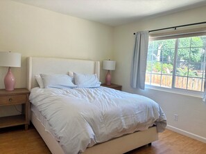 Cama bem arrumada com colcha listrada, duas luminárias de cabeceira e janela com vista para vegetação.