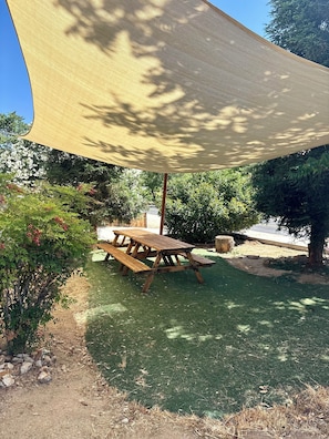 A wooden picnic table under a large shade sail, surrounded by greenery and trees.