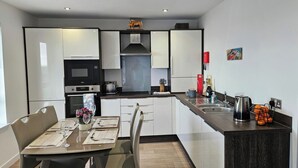 Une cuisine moderne avec des meubles blancs, un coin repas avec une table dressée pour deux personnes, et un petit îlot central avec une bouilloire et des fruits, notamment des oranges.