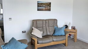 Un salon avec un canapé en cuir, deux coussins bleus, une petite table d’appoint en bois sur laquelle se trouve un objet blanc, et un tableau accroché au mur.