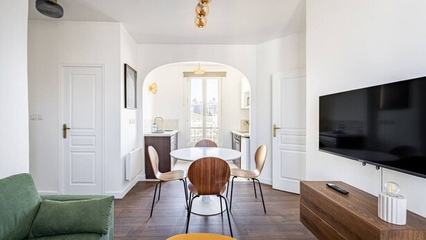 En moderne stue med et fladskærms-tv, en sofa, et spisebord med stole, og et køkkenområde kan ses gennem en bue.