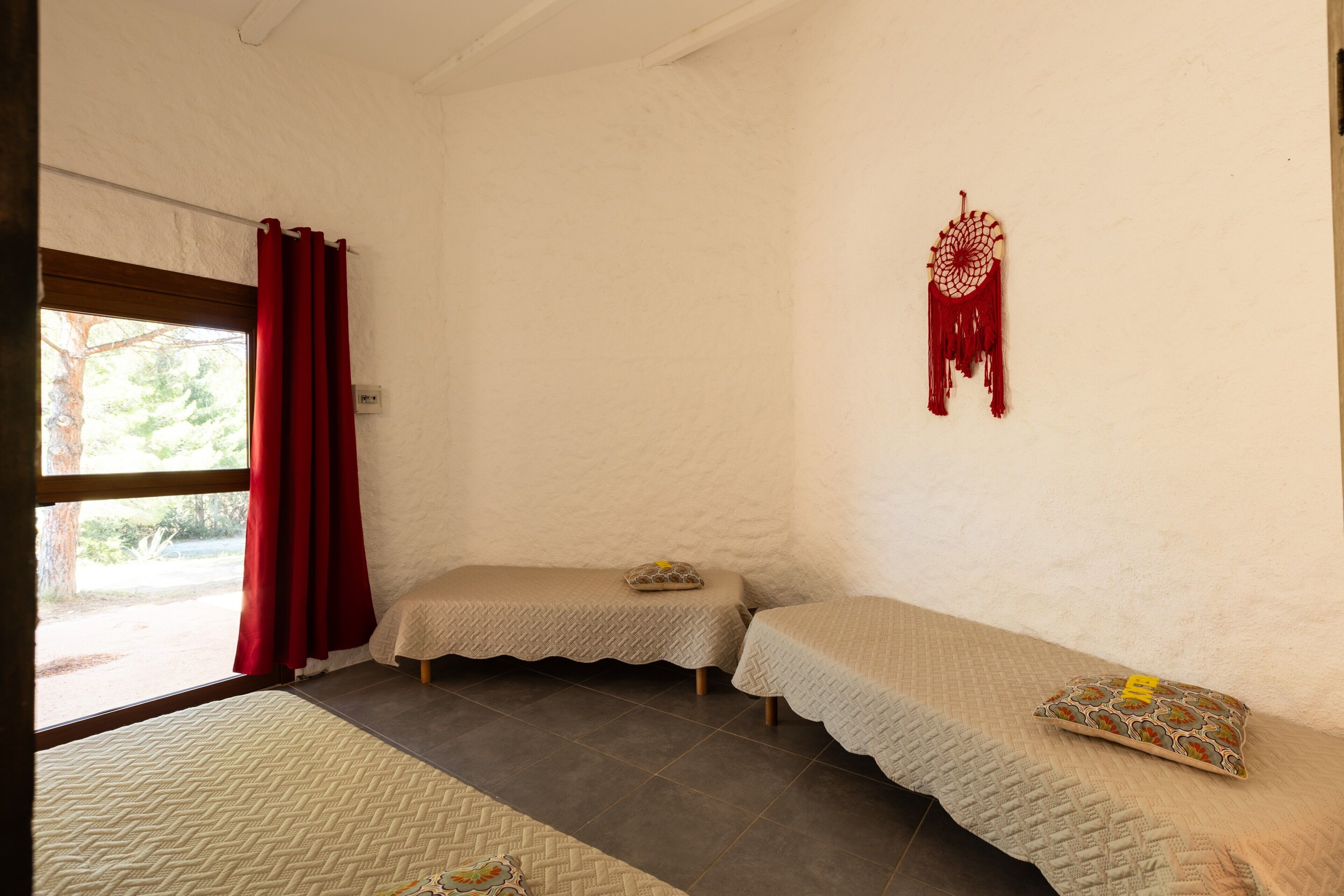 A room with two beds, a window with red curtains, a dreamcatcher, and a view of greenery outside.