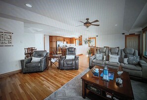 A spacious living room with a ceiling fan, a fireplace, and a kitchen in the background.