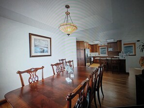 A dining room with a wooden table and chairs, a chandelier, and a kitchen in the background.