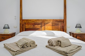 Habitación de hotel con cabecera de madera, dos mesitas de noche con lámparas, cama bien hecha con toallas enrolladas y una pared blanca.