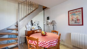 Un comedor con mantel a cuadros, sillas de madera, un frutero, una escalera, un radiador y un cartel antiguo en la pared.