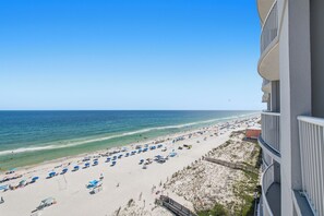 A beach view from a balcony with a clear sky, a sandy beach with umbrellas, and a building with balconies. - Corner Condo - 3 bedroom/3 bath - Beautiful direct view of Gulf of America (Gulf Shores)