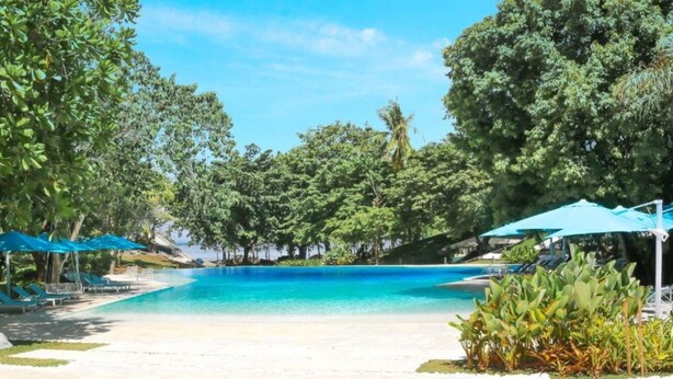 Ein klares, blaues Schwimmbecken, umgeben von üppiger Vegetation, mit Liegestühlen und Sonnenschirmen.