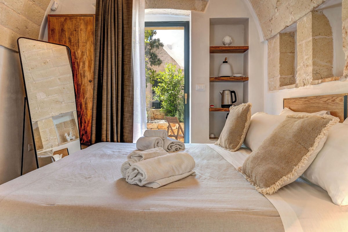 A bedroom with a bed, towels, a wooden headboard, a shelf with a kettle, and a view of the outdoors.