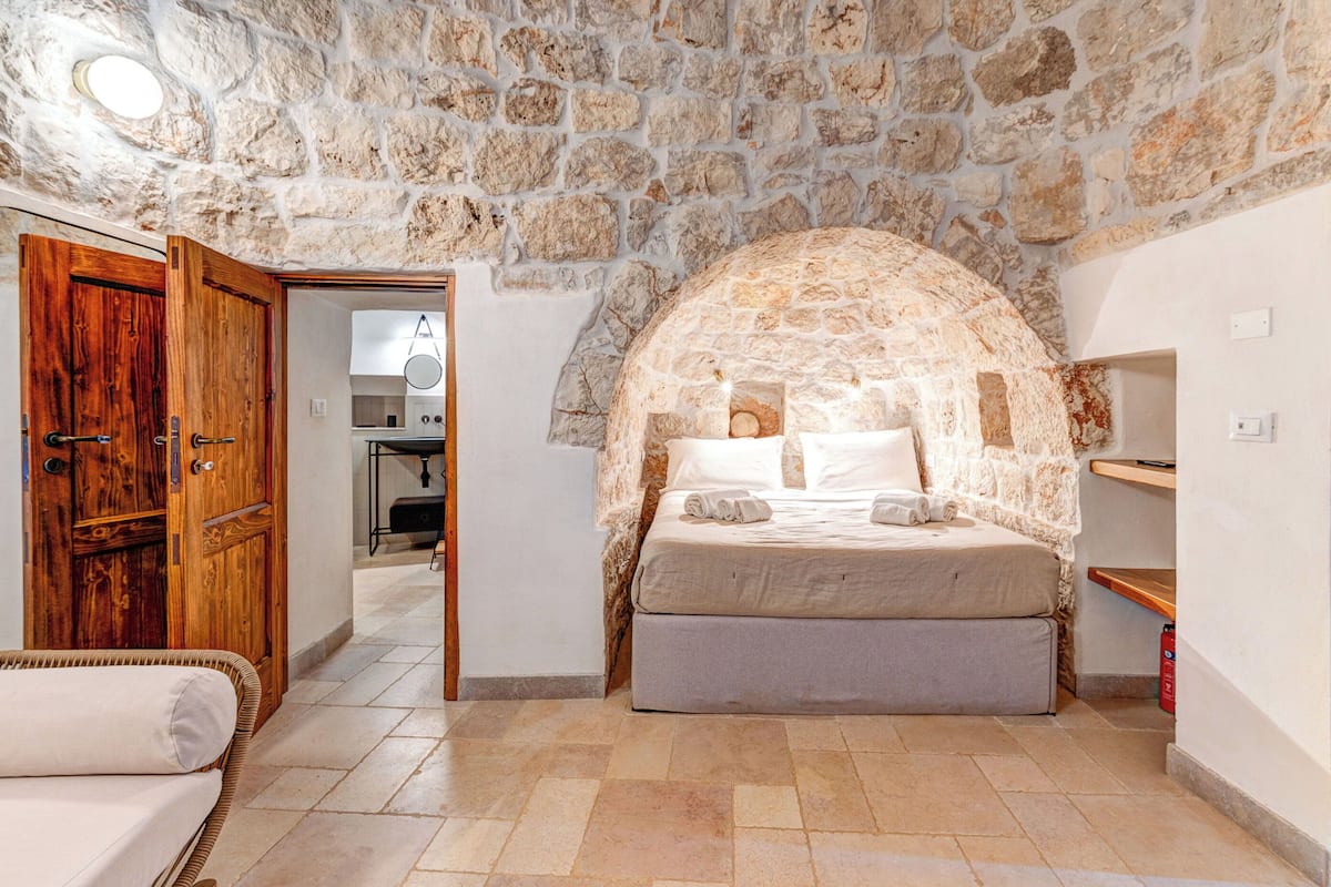 A stone-walled bedroom with a bed, a wooden door, and a kitchenette in the background.