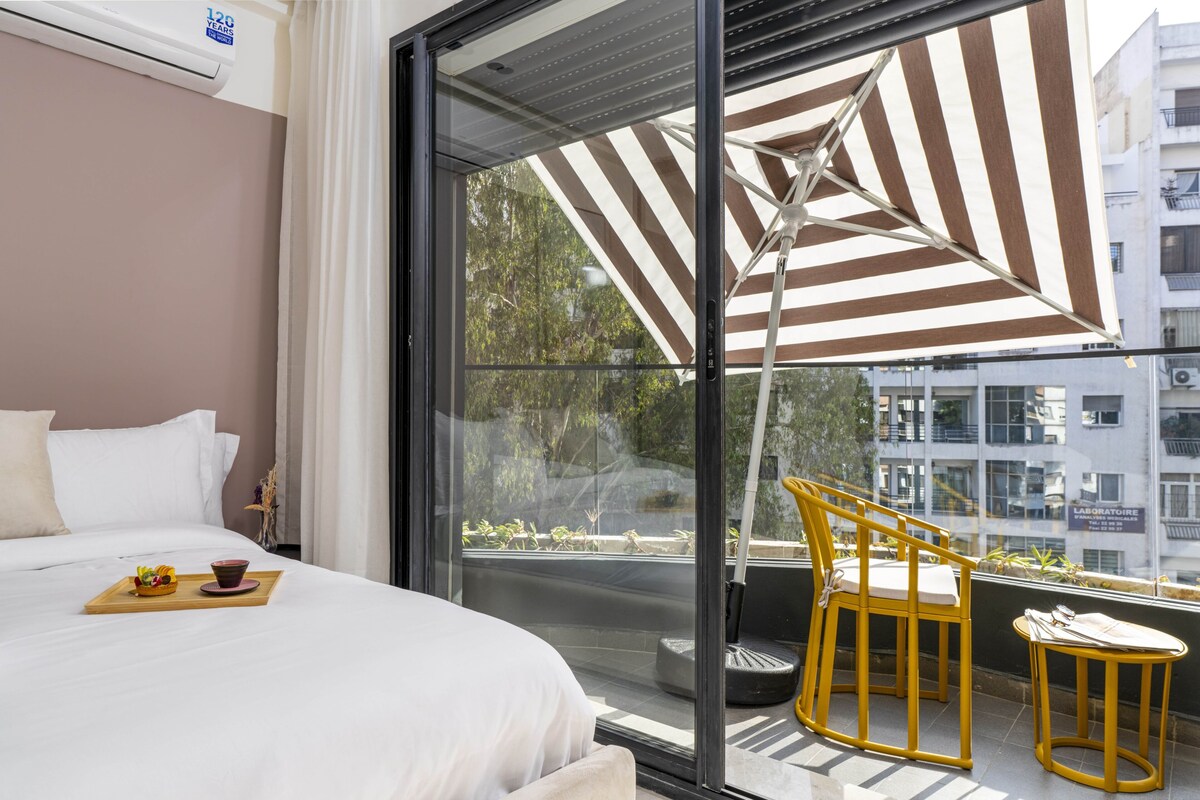 Habitación de hotel con cama, zona de comedor con una bandeja de comida, una silla y un amplio ventanal con vistas a edificios y zonas verdes.