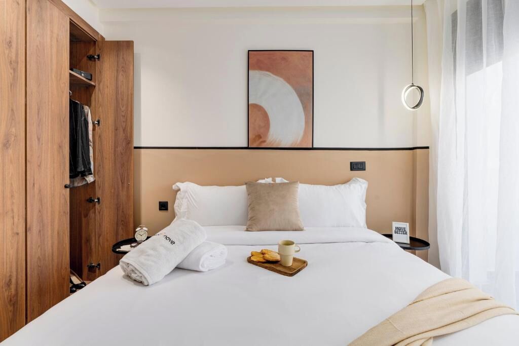 Une chambre d’hôtel avec un lit, une armoire en bois, un tableau, une table de chevet, une horloge et un plateau avec des biscuits et une tasse.