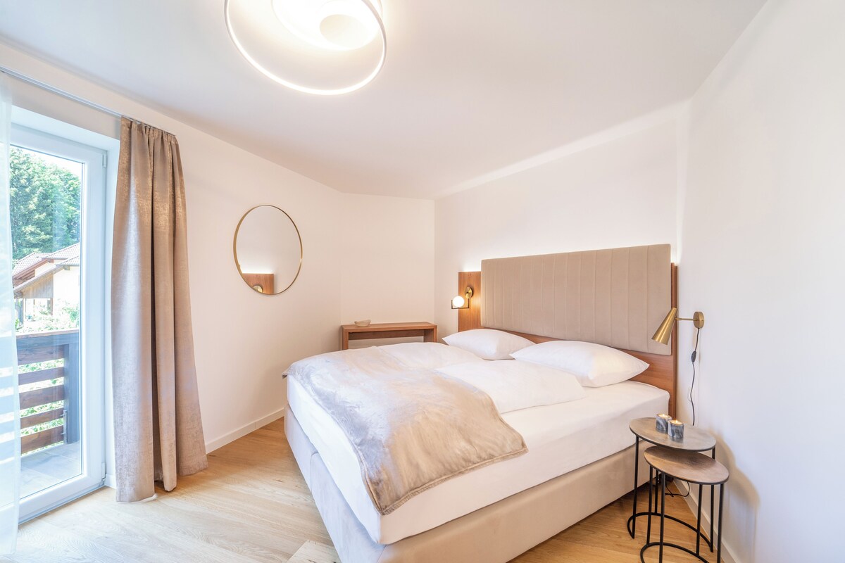 A modern bedroom with a large bed, a bedside table, a round mirror, and a small round table with a lamp.