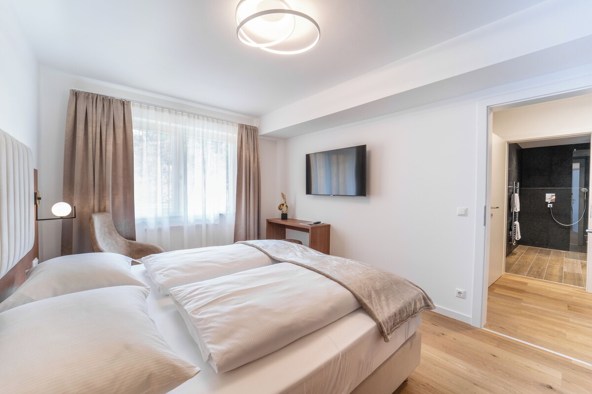 A modern bedroom with a large bed, a bedside table, a flat-screen TV, and a shower area.