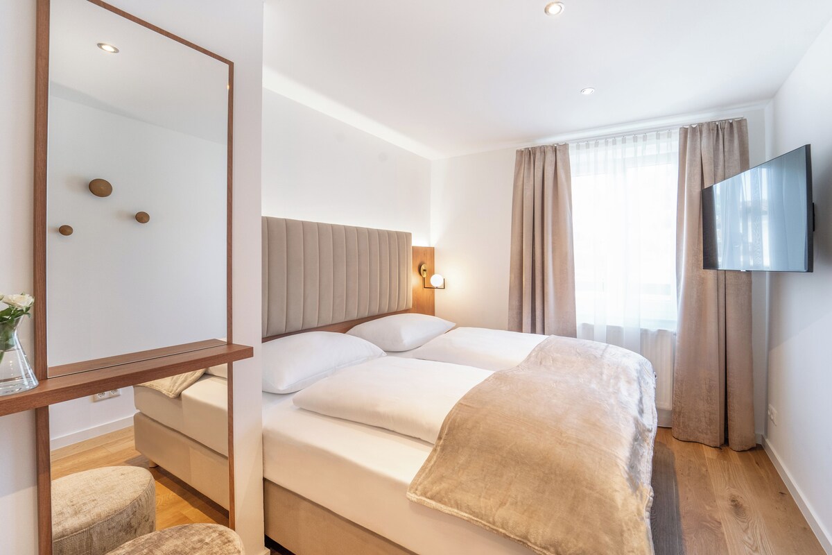 A hotel room with a bed, a bedside table, a mirror, a TV, and a window with curtains.