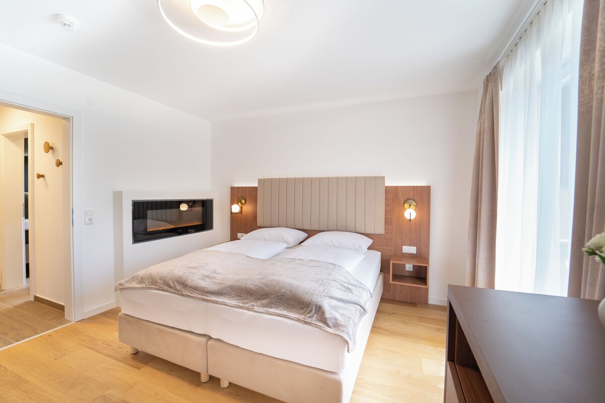 A modern bedroom with a large bed, bedside tables, a TV mounted on the wall, and a window with curtains.