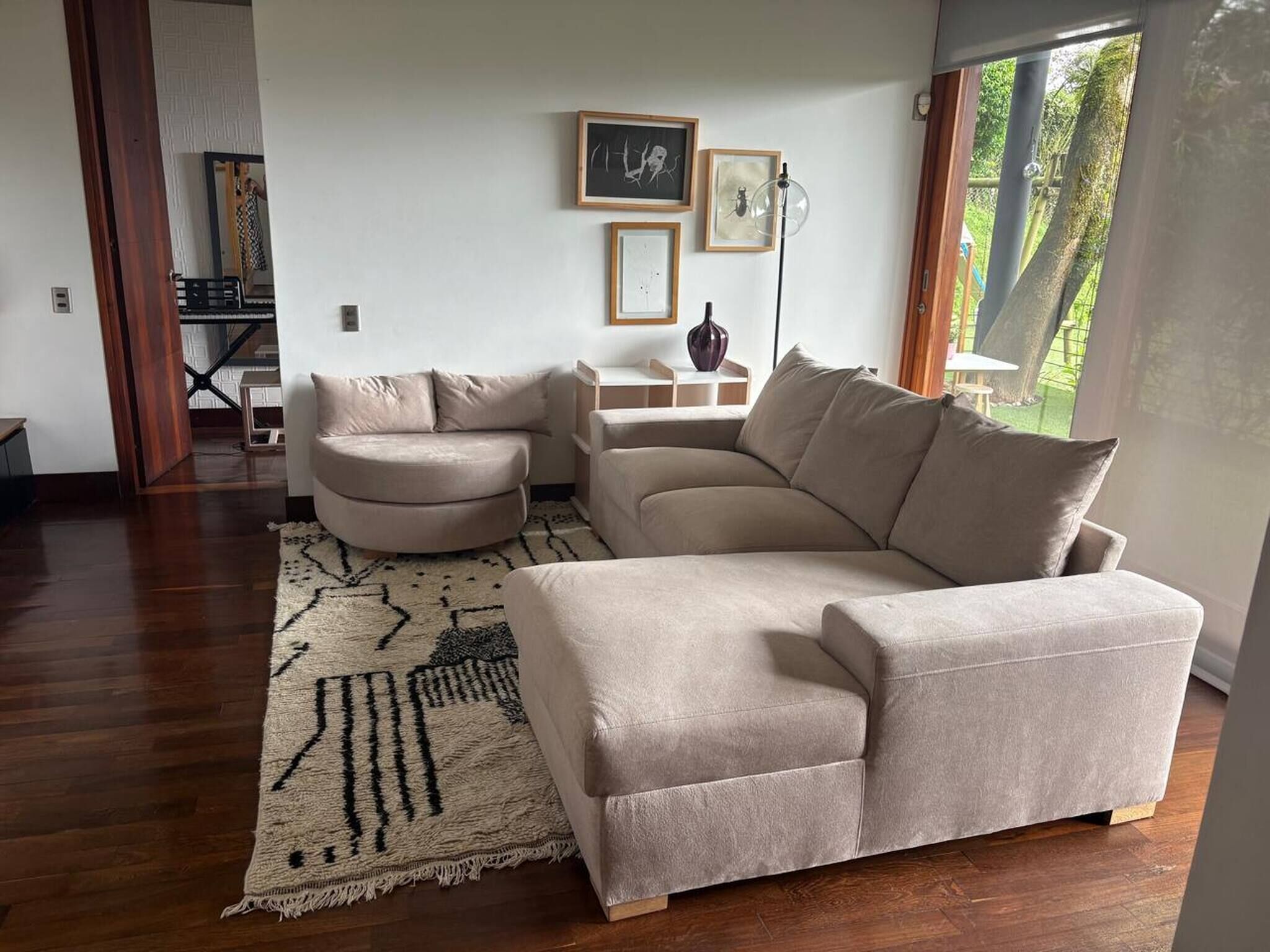 A living room with a beige sectional sofa, a round ottoman, a patterned rug, and a small side table with a vase.