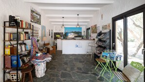 A room with a stone floor, a reception desk, bookshelves, a wicker chair, and a view through a large window. - Mobile Home 'Village Camping De L'ostriconi 27' with Sea View, Shared Pool and Wi-Fi (Palasca)