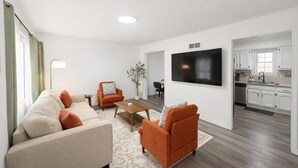 A modern living room with a sofa, armchairs, a coffee table, and a TV. There is a kitchen area with white cabinets and a window. - 4BR Beautiful Gardner Getaway Game Room Central Location Sleeps 10 (Gardner)