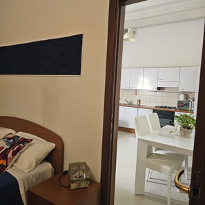A room with a bed, a bedside table, a small table with a chair, and a view into a kitchen with white cabinets and a stove. - The walls of lovers (Verona)