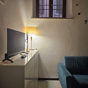A room with a TV on a white desk, a lamp, a blue couch, and a window with a view of buildings. - The walls of lovers (Verona)
