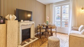 A modern living room with a fireplace, a TV, a dining table with chairs, and a large window with a view of buildings. - Stunning apartment with AC - Center of Paris (Paris)