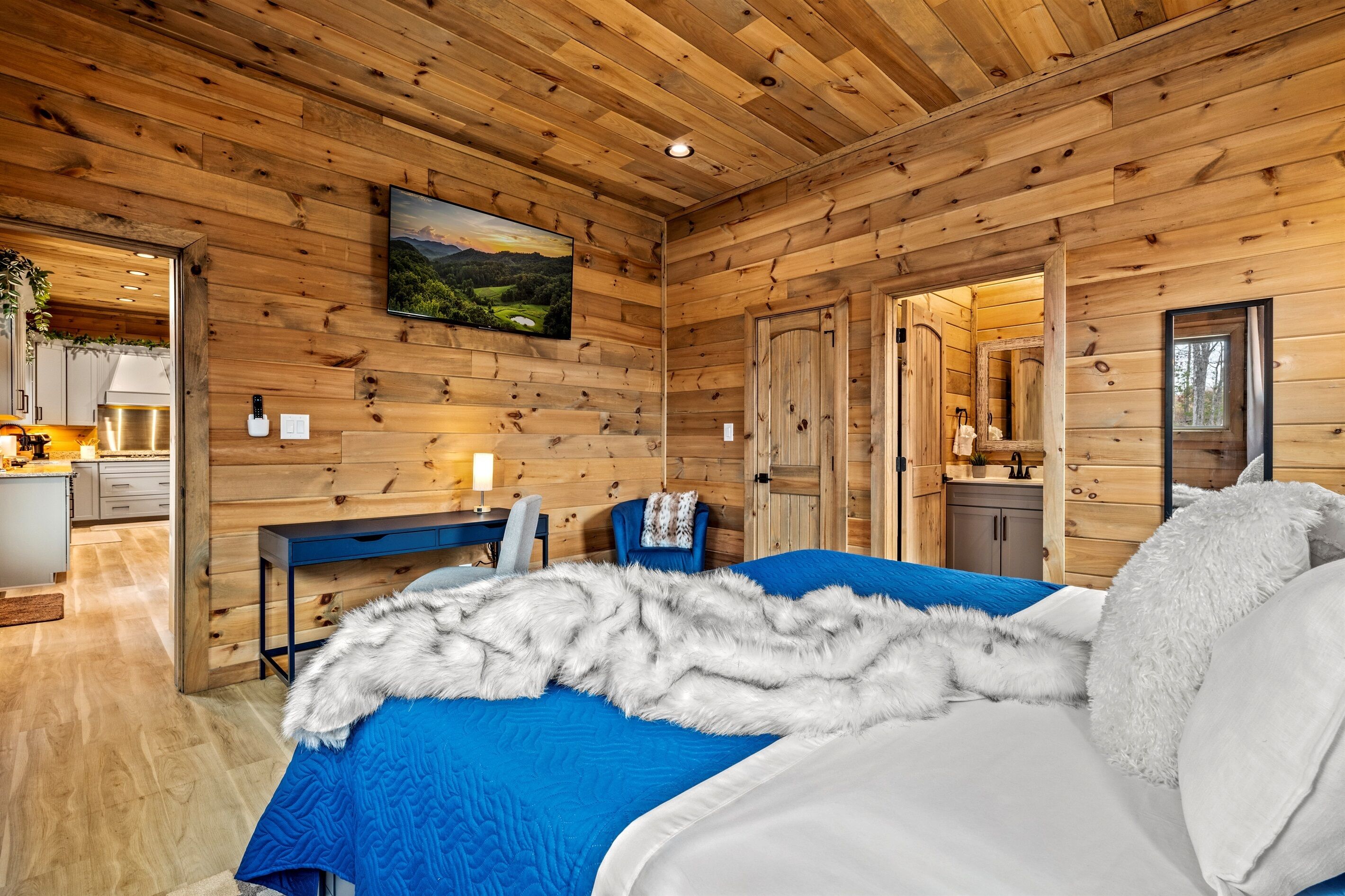 A wooden-paneled room with a bed, a desk, and a view of a kitchen and bathroom through an open door.