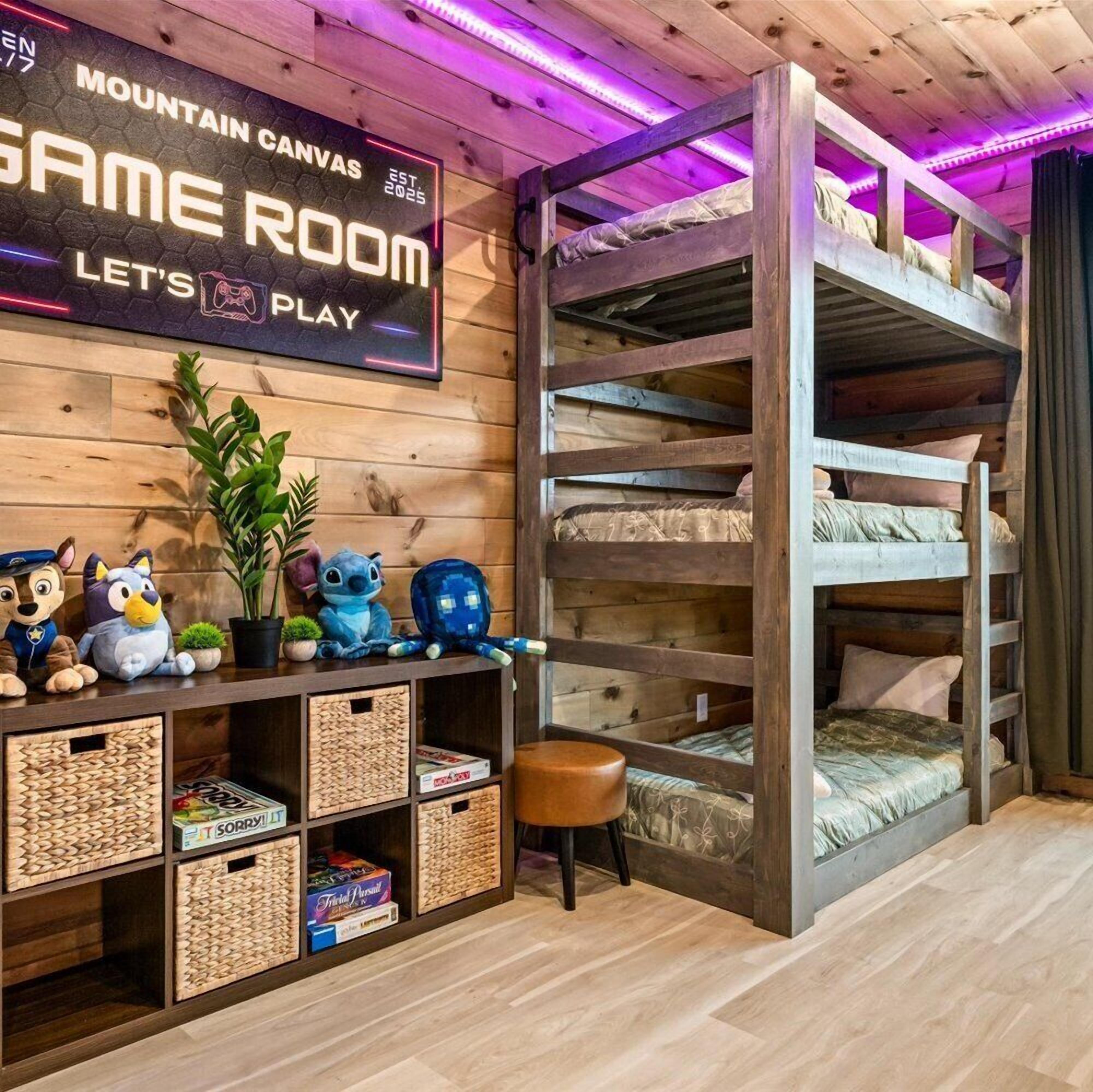 Et hyggeligt værelse med køjesenge, en hylde med bløde legetøj, og et skilt der står 'MOUNTAIN CANVAS GAME ROOM LET'S PLAY'.
