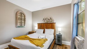 A bedroom with a wooden bed, white bedding, a yellow throw blanket, a decorative wall piece, a nightstand with a lamp, and a window with a curtain. - 3BR near Downtown COS with fenced Backyard Seating (Colorado Springs)