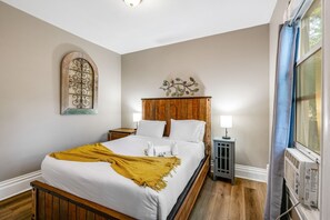 A bedroom with a wooden bed, white bedding, a yellow throw blanket, a decorative wall piece, a nightstand with a lamp, and a window with a curtain. - 3BR near Downtown COS with fenced Backyard Seating (Colorado Springs)