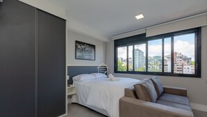 Une chambre moderne avec un lit, une table de chevet, un canapé, une fenêtre donnant sur la ville et un tableau encadré au mur.