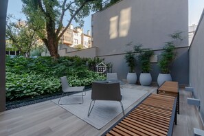 Eine Terrasse im Freien mit einer Holzbank, einem Tisch und Stühlen, umgeben von üppigem Grün und Topfpflanzen.