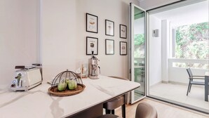 Cozinha moderna com bancada branca, máquina de café e torradeira. Quadros emoldurados na parede e vista para o exterior por uma porta de vidro.