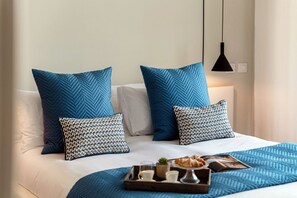 Ein ordentlich bezogenes Bett mit blau-weißen Kissen, ein Tablett mit einem Croissant, eine kleine Pflanze und eine Zeitschrift auf einer blauen Bettdecke.