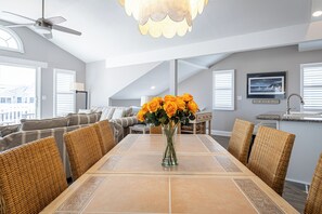 Un comedor con una mesa, sillas, un jarrón de flores, un ventilador de techo y una cocina al fondo.